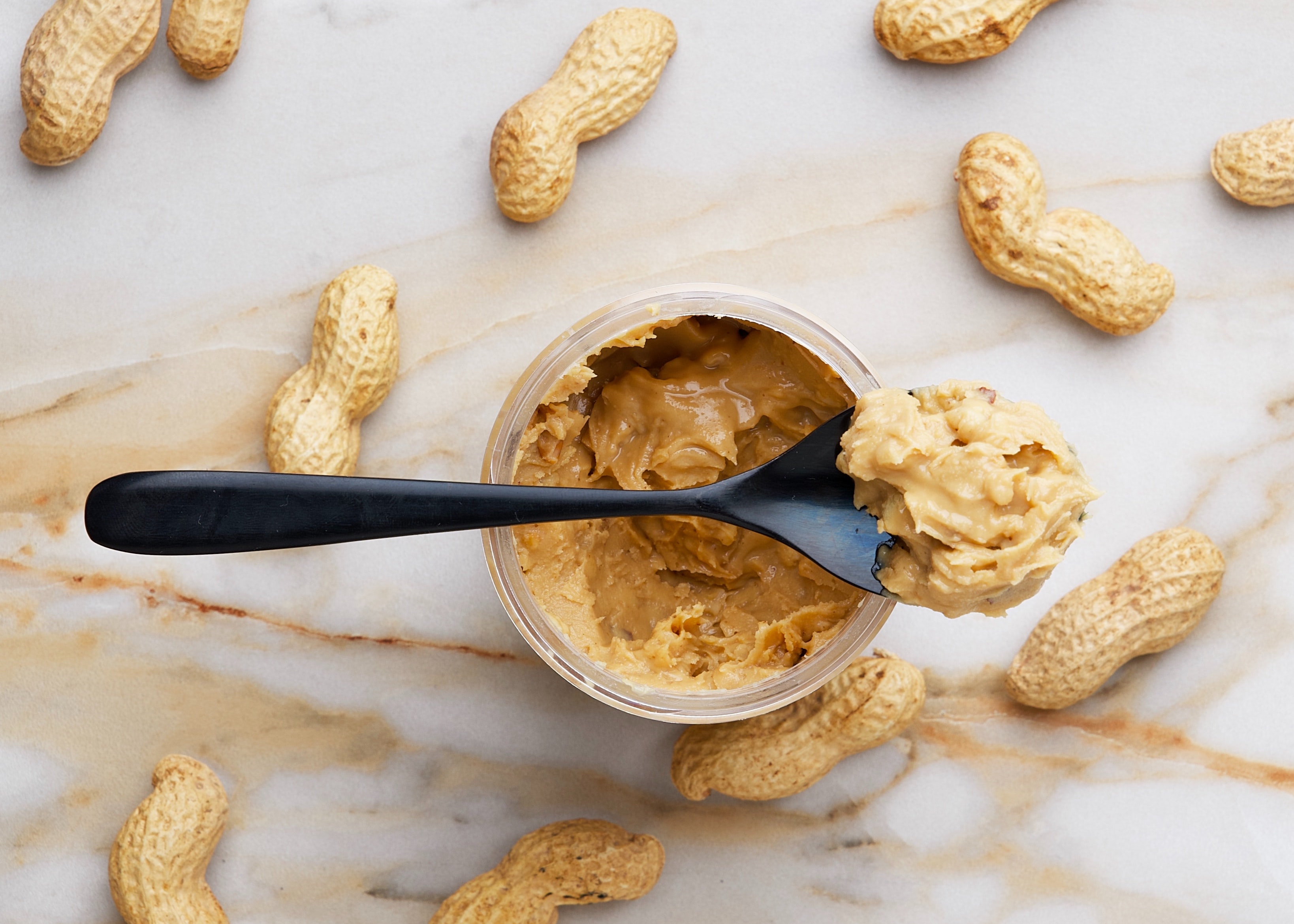 Pot de beurre de cacahuètes avec une cueillière, et des cacahuètes éparpillées autour